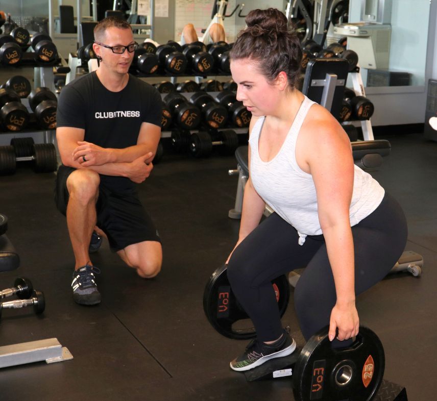 Coach Donovan watching closely as a young client performs squats on heel wedges with weight plates to target the quads during a gym training session.