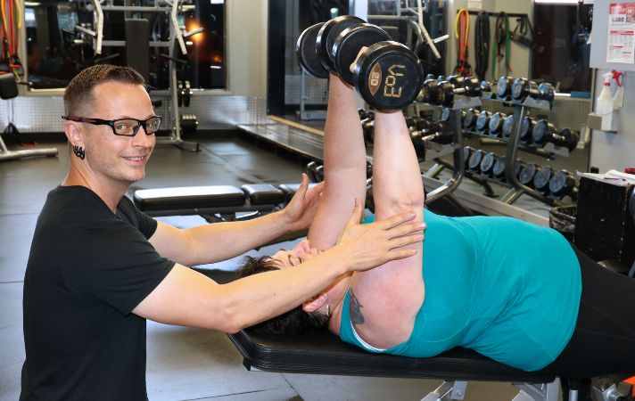 Coach Donovan spotting an overweight middle-aged female client performing a dumbbell bench press during personal training session.