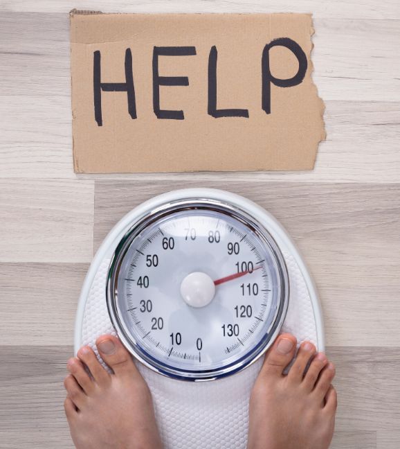 Overweight person standing on a bathroom scale with a ‘Help’ sign overhead — symbolizing the need for a personal trainer and weight loss support.